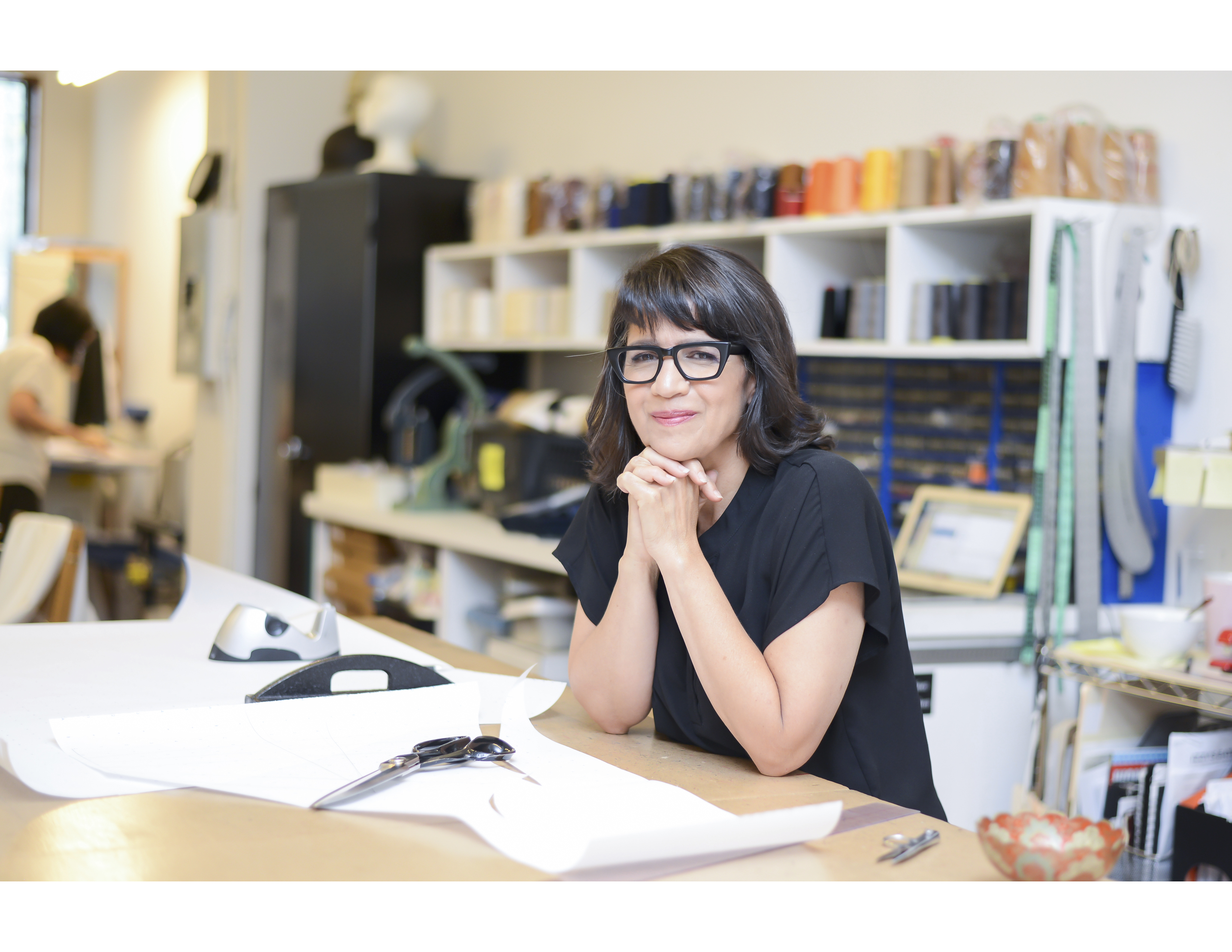 woman with short dark hair and glasses leans on a large table surrounded by sewing and drawing tools